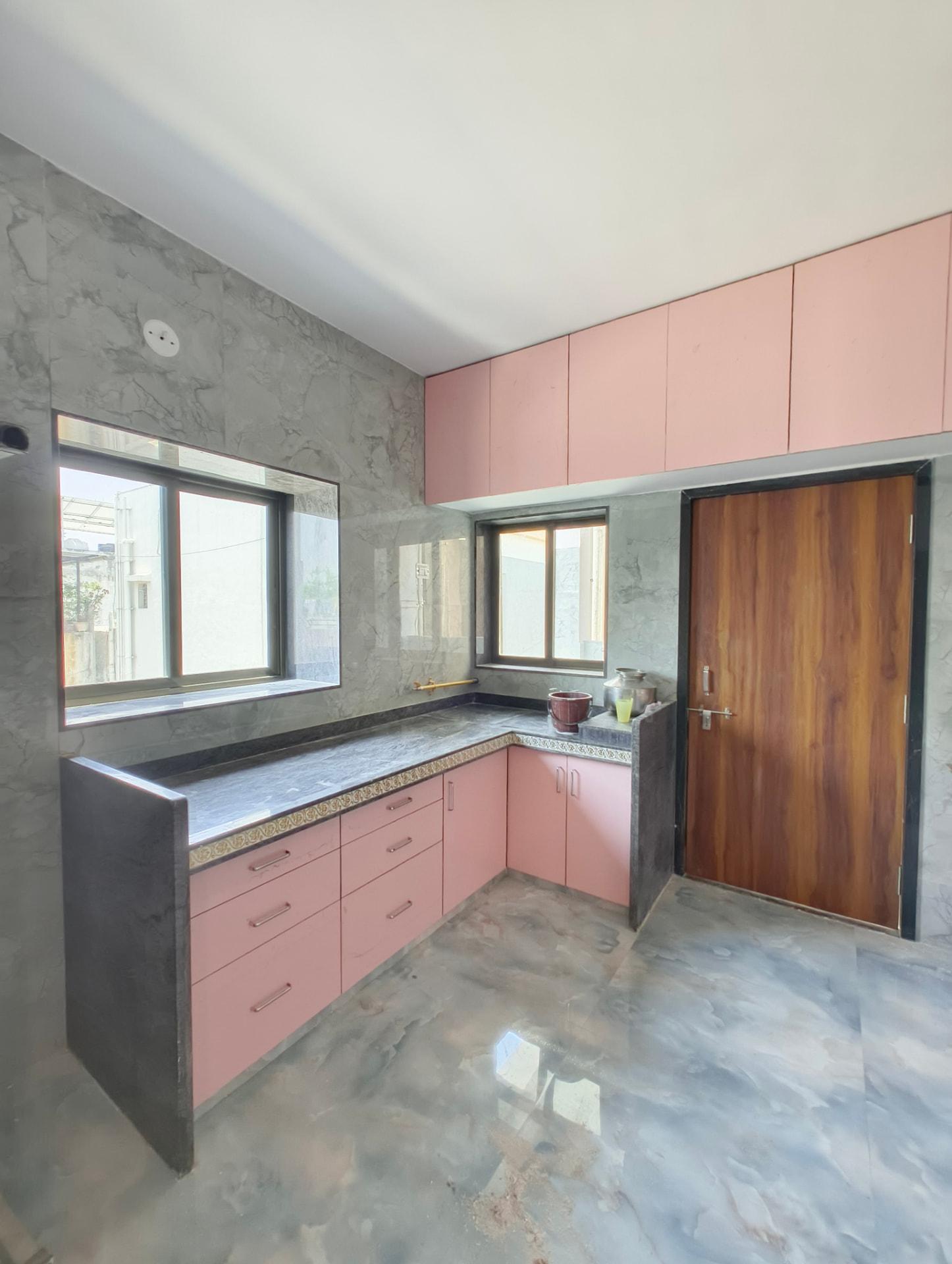 L-shaped blush pink modular kitchen with dark countertop, marble-look splashback and a solid wooden entry door